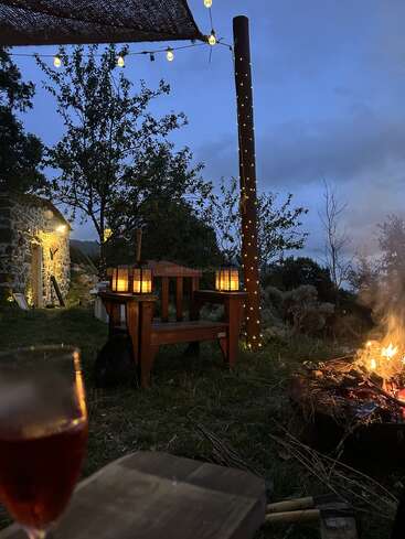 Cozy outdoor scene at dusk with a glowing fire pit, string lights, rustic stone house, wooden chairs, lanterns, and a glass of red drink in foreground.