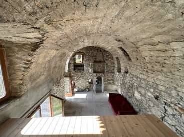 This image shows a rustic stone room with arched ceilings, exposed masonry, two windows, a red sofa, wooden floor, and abundant natural light streaming in.