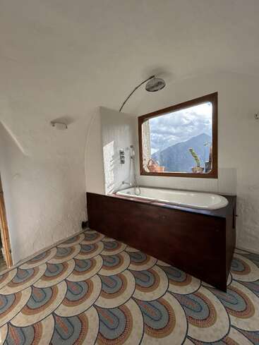 A bathroom with a bathtub and shower, patterned floor tiles, white textured walls, and a large window offering a scenic mountain view with potted plants outside.