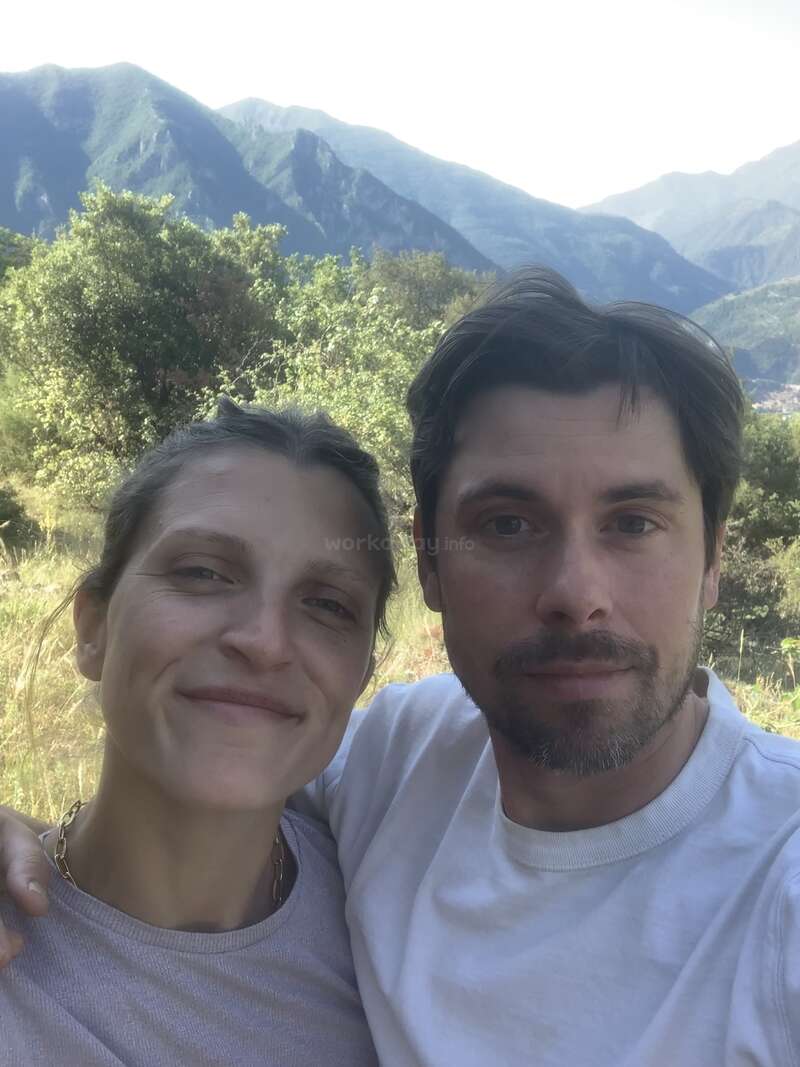The image shows a selfie of a man and woman in front of a mountainous landscape with trees and grass, wearing casual clothing.
