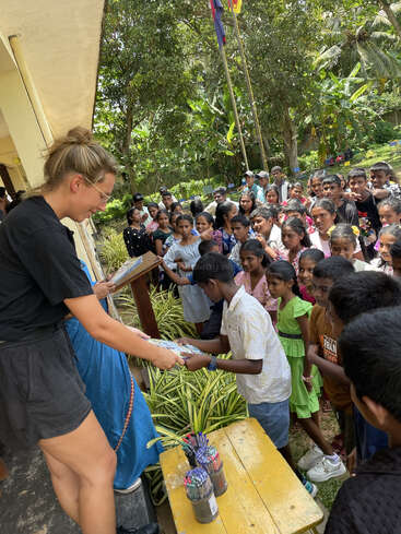 Une femme distribue des fournitures scolaires à un groupe d'enfants en plein air, tandis que les enfants attendent impatiemment dans la file d'attente. L'atmosphère est joyeuse et axée sur la communauté.
