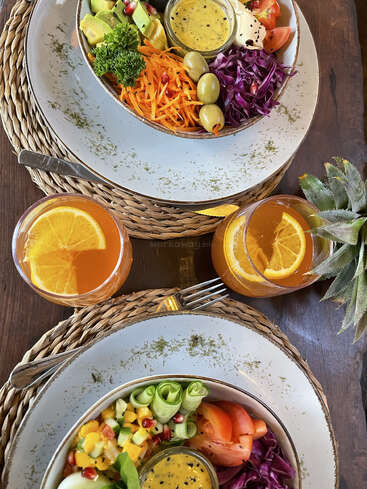 Deux salades vibrantes servies dans des bols avec des légumes colorés, de la vinaigrette et des garnitures de fruits. Accompagnées de deux verres de boisson à l'orange, joliment présentées sur des sets de table rustiques.