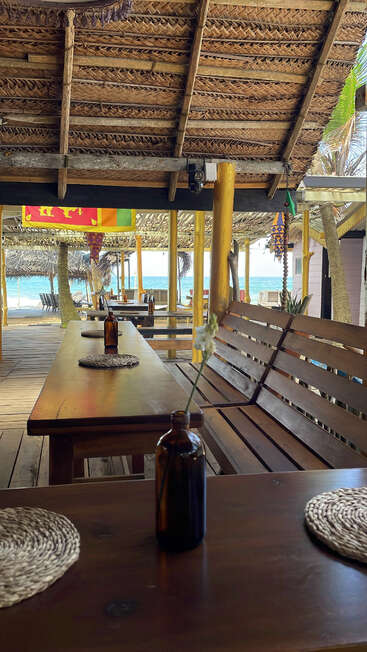 Un restaurant rustique en bord de mer avec des tables en bois, des bancs et des sets de table tressés. Des bouteilles en verre servent de vases. Toit de chaume, décor tropical, vue sur l'océan et drapeaux éclatants.