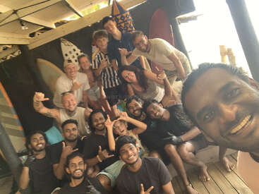 Un groupe de personnes joyeuses pose ensemble à l'intérieur près de planches de surf, certaines faisant des signes de la main en forme de shaka. L'atmosphère est détendue, amicale, pleine de sourires et de rires.