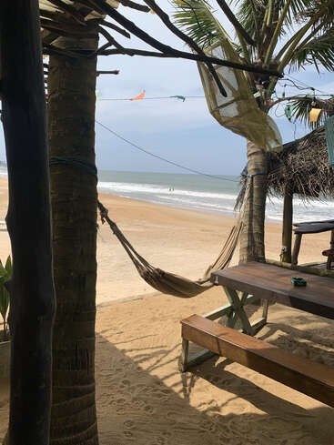 Une scène de plage tranquille avec un hamac, une table en bois, des bancs, des palmiers et des drapeaux colorés, le tout surplombant le sable doré et les vagues de l'océan qui roulent doucement. Atmosphère paisible.