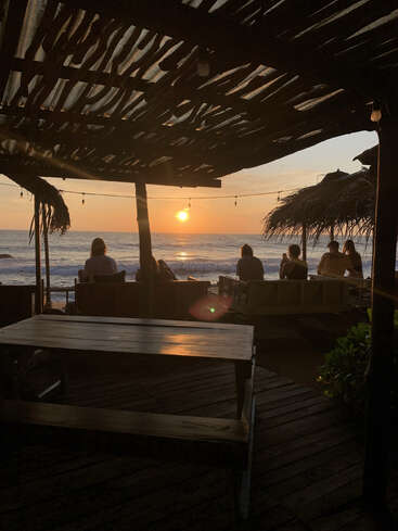 Un groupe de personnes est assis sur des bancs sous un abri rustique, regardant le coucher de soleil sur l'océan. La lumière chaude crée une atmosphère paisible et relaxante.