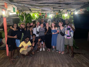 Un groupe d'amis pose ensemble la nuit, souriant et faisant des signes de paix, debout sur une terrasse en bois décorée de plantes et de lumières colorées, créant une atmosphère festive.