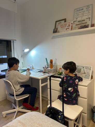 Two children sit at adjacent desks, drawing or doing homework. The room is cozy, well-lit, with diplomas and awards displayed on a shelf above them.