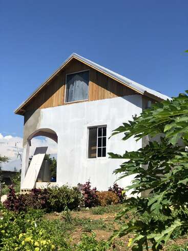 The image depicts a white house with a gable roof, featuring a wooden upper section and a window, surrounded by lush greenery and set against a clear blue sky.
