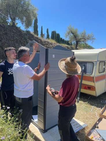 Three people are assembling an outdoor shed under a clear blue sky, near a camper. One uses a power drill; wildflowers and trees surround the sunny scene.