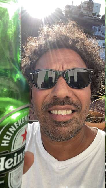 Ein Mann mit Sonnenbrille und weißem Hemd lächelt und hält eine Heineken-Bierflasche in der Hand. Das Sonnenlicht scheint hinter ihm und spiegelt sich in seiner Sonnenbrille. Entspannte, lockere Atmosphäre.