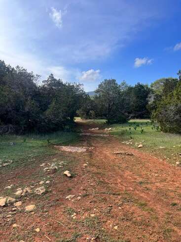 Ein gewundener Feldweg führt durch eine grüne Landschaft, die von Bäumen gesäumt ist. Der Himmel über uns ist strahlend blau mit vereinzelten Wolken, die an Ruhe und Abenteuer erinnern.