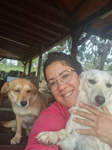 Une femme souriante, portant des lunettes, serre dans ses bras un chien blanc, tandis qu'un autre chien, de couleur fauve, est assis à proximité sur une terrasse en bois, avec des arbres et de la verdure en arrière-plan.