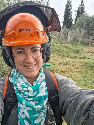 Une personne souriante, coiffée d'un casque de sécurité orange, portant des casques antibruit, des lunettes et un foulard bleu à motifs floraux, se tient à l'extérieur sur une pelouse, avec des arbres en arrière-plan.
