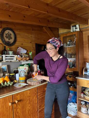 Dans une cuisine chaleureuse en bois, une femme portant des lunettes et un foulard coloré coupe joyeusement une orange sur une planche à découper, entourée de produits alimentaires.