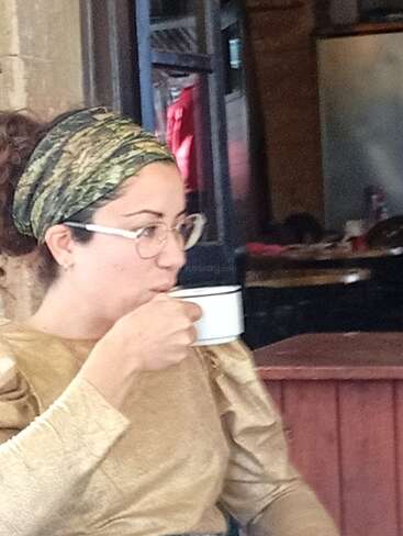 Une femme portant des lunettes et un foulard vert boit une gorgée dans une tasse blanche. Elle porte un haut doré et est assise à l'intérieur d'un café chaleureux.
