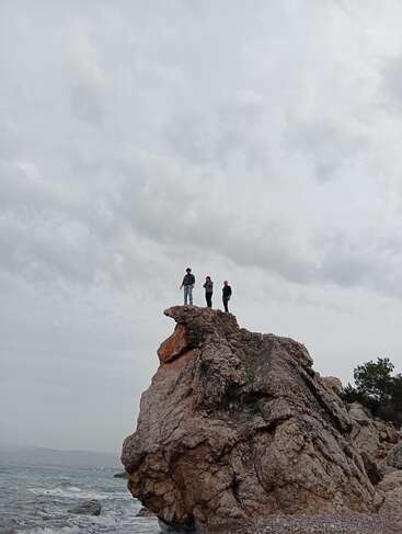Trois personnes se tiennent au sommet d'une grande falaise rocheuse au bord de la mer, sous un ciel nuageux, le regard tourné vers l'eau, créant ainsi une scène spectaculaire et pleine d'aventure.