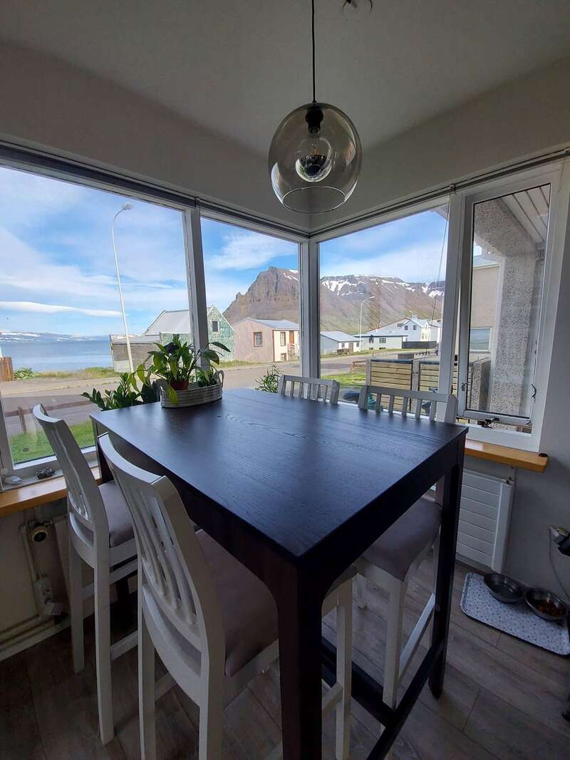 La imagen representa un comedor con una mesa y sillas, provisto de un gran ventanal con vistas al océano y a un paisaje montañoso.