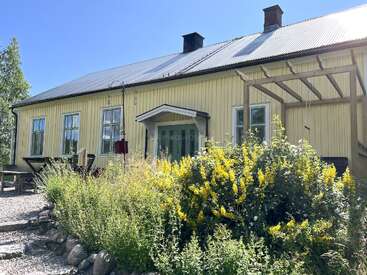 A yellow wooden house with a metal roof, surrounded by vibrant green plants and yellow flowers, sits under a clear blue sky on a sunny day.