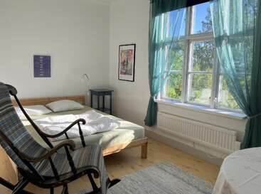 A bright, cozy bedroom with a wooden bed, rocking chair, blue curtains, large window, paintings on white walls, and sunlight streaming onto a wooden floor.