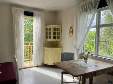 Sunlit room with white curtains, wooden floors, and vintage furniture. A yellow corner cabinet, rustic table, and flowers create a cozy, inviting, peaceful atmosphere with greenery outside.