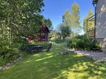 A peaceful garden scene with lush green grass, outdoor seating, stone-bordered flowerbeds, a red barn, tall trees, and a clear blue sky create a serene atmosphere.