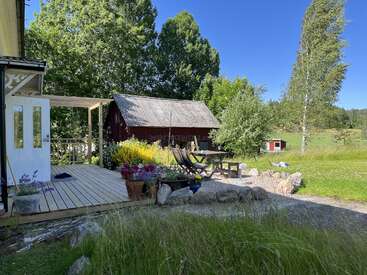 A cozy countryside scene featuring a wooden deck with potted flowers, outdoor chairs, a rustic red barn, lush green trees, and a bright blue sky.