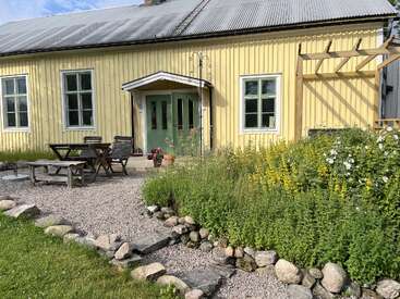 Una encantadora casa de madera amarilla con puertas verdes, rodeada de exuberantes plantas en flor, caminos de grava, rústicos asientos al aire libre y un borde de piedra bajo un cielo soleado.