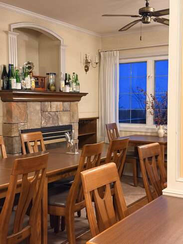 A cozy dining room with wooden tables and chairs, stone fireplace with bottles and decor, ceiling fan, large window, white curtains, and warm lighting.