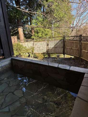 A tranquil outdoor hot spring bath with stone edges, surrounded by bamboo fencing, trees, and greenery, under a clear blue sky. Peaceful and inviting atmosphere.