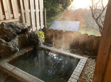 A serene outdoor onsen with steaming hot water, natural stones, surrounded by wooden fencing, overlooks a tranquil landscape of autumn trees and a rustic shed. Peaceful atmosphere.