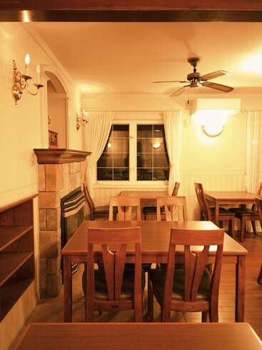 A cozy, warmly-lit dining room with wooden tables and chairs, stone fireplace, ceiling fan, white curtains, wall lights, and wooden shelving, creating a welcoming atmosphere.