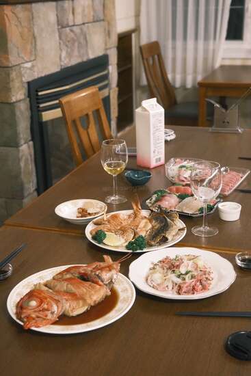A cozy dining setting features plates of cooked and raw fish, salads, wine glasses, chopsticks, and a milk carton on a wooden table near a fireplace.