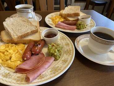Two breakfast plates with scrambled eggs, ham, sausages, guacamole, toast, and jam. Two cups of black coffee sit beside the plates on a wooden table.