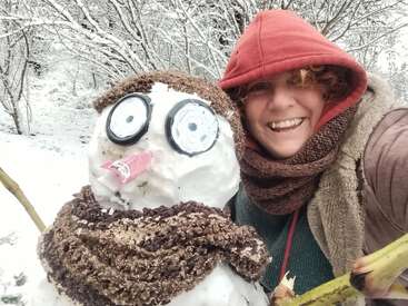 A smiling person in a red hood poses with a whimsical snowman, both bundled in brown scarves, surrounded by snowy trees in a winter wonderland scene.
