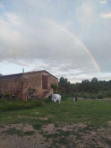 A rustic stone house sits surrounded by grass and nature. Above, a faint rainbow arcs across a cloudy sky, adding a magical touch to the peaceful scene.