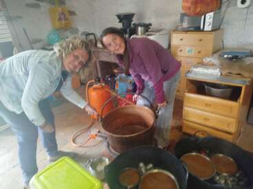 Two women are smiling while cooking together in a rustic kitchen. They are working with a large copper pot and other kitchenware, possibly preparing traditional food.