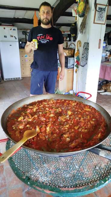 A man stands indoors holding a drink, behind a large pan of tomato-based stew on a green table. Cozy kitchen, relaxed atmosphere, homey decorations visible.