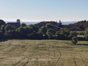 A peaceful rural scene with a sunlit field, old stone buildings, trees, and power lines. Mountains appear in the background beneath a clear sky.