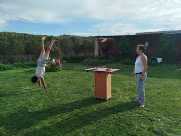 A person does a handstand on green grass while another stands nearby watching. There’s a small table with objects on top, set in a scenic outdoor area.
