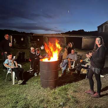 Eine Gruppe von Leuten versammelt sich nachts draußen um ein Fassfeuer, sitzt auf Stühlen, unterhält sich und genießt die Wärme unter dem dunklen Abendhimmel.
