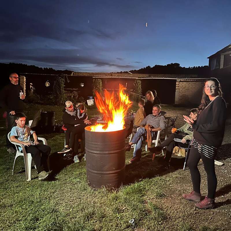 A group of people gather around a barrel fire outdoors at night, sitting on chairs, chatting, and enjoying the warmth under a dark, twilight sky.