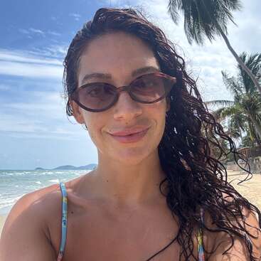 Una mujer con el pelo mojado y rizado y gafas de sol sonríe a la cámara en una playa soleada. Al fondo se ven palmeras, el cielo azul y las olas del mar.