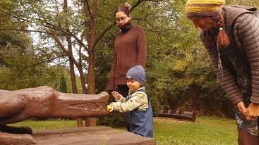 Uma criança pequena com um chapéu azul toca a boca de uma escultura de crocodilo, observada por duas mulheres vestidas com roupas quentes, em meio a árvores verdes e grama.