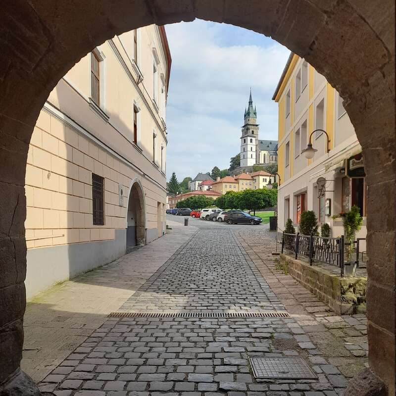 Uma pitoresca rua de paralelepípedos, emoldurada por um arco de pedra e ladeada por prédios em tons pastéis, sobe em direção a uma charmosa torre de igreja sob um céu parcialmente nublado.
