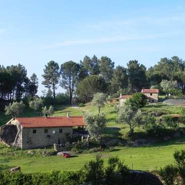 L'image représente un paysage rural serein, avec une maison en pierre au toit rouge, entourée d'une végétation luxuriante et d'arbres, le tout dans un ciel bleu limpide.