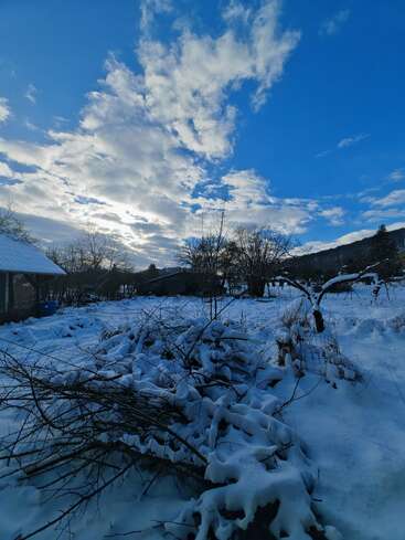 Snow blankets a rural landscape with bare trees and a small house, while dramatic clouds and blue sky create a serene, cold winter atmosphere in the countryside.