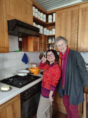 Two women smile in a cozy kitchen. One gives a peace sign, the other stands beside her. Wooden cabinets and labeled jars decorate the background warmly.