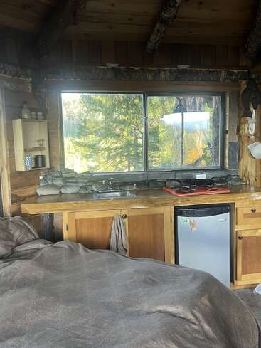 El interior de una acogedora cabaña rústica cuenta con una cocina de madera, una mininevera y una gran ventana con vistas al bosque. Cama con fundas marrones en primer plano. Ambiente tranquilo.