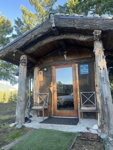 Una cabaña rústica de madera con una puerta de cristal, astas sobre la entrada, dos sillas, rodeada de árboles altos. Un letrero humorístico dice: "HIPPIES USE BACKDOOR".